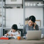 Young Asian mother working from home on a laptop and talking on the phone while little daughter is studying from home. She is attending online school classes with a digital tablet and doing homework at home